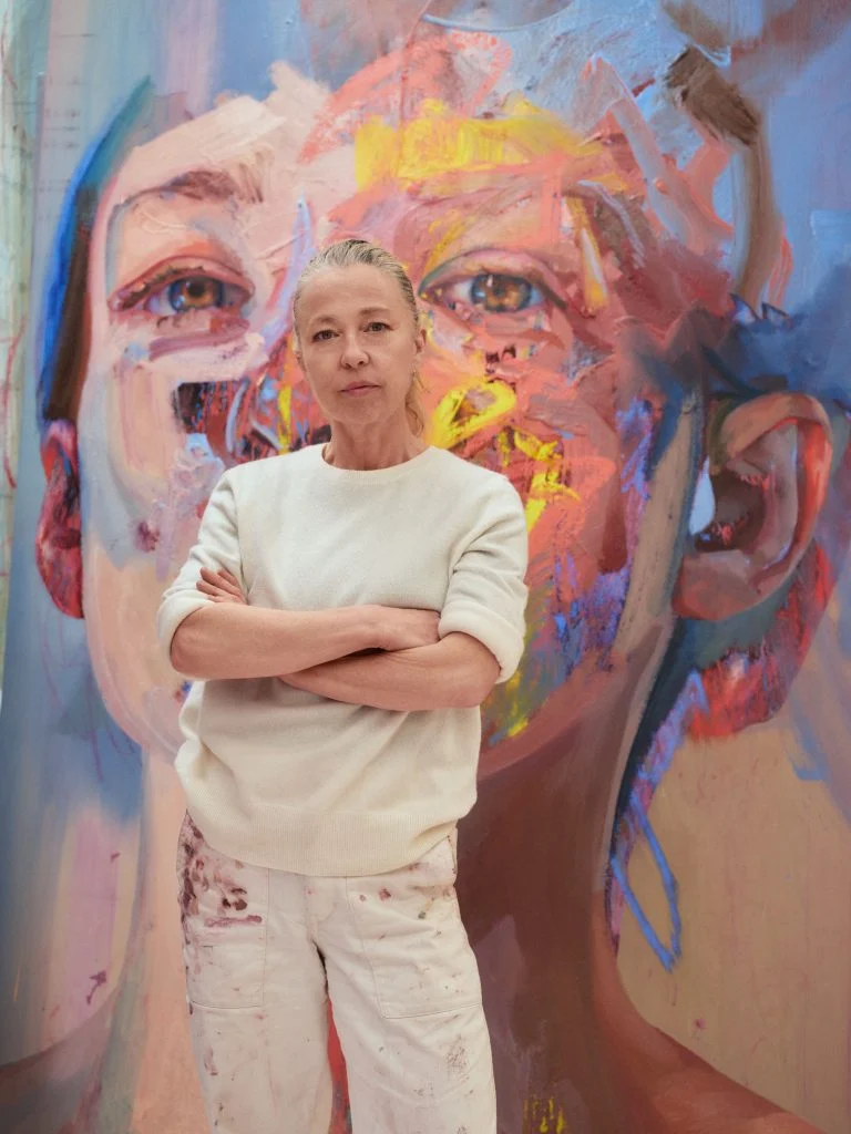 A woman in white sweater and paint-splattered paints standing in front of a portrait